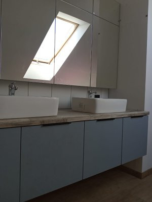 Łazienka z niebieskimi szafkami pod umywalkami, drewnianym blatem i lustrami odbijającymi okno dachowe.