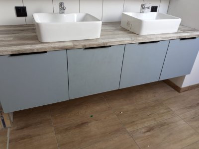 Łazienkowa szafka podumywalkowa w odcieniach szarości z dwoma białymi umywalkami nablatowymi, czarne minimalistyczne uchwyty, blat imitujący drewno, płytki ceramiczne na ścianie.