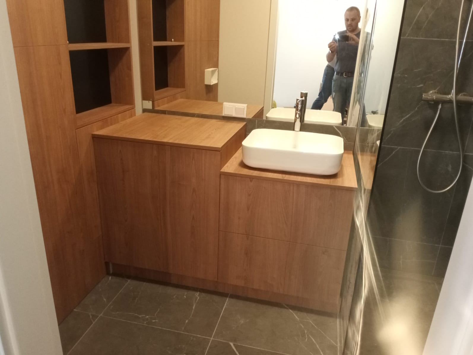 Drewniana zabudowa łazienkowa z szafkami i umywalką nablatową; w lustrze odbija się osoba robiąca zdjęcie.