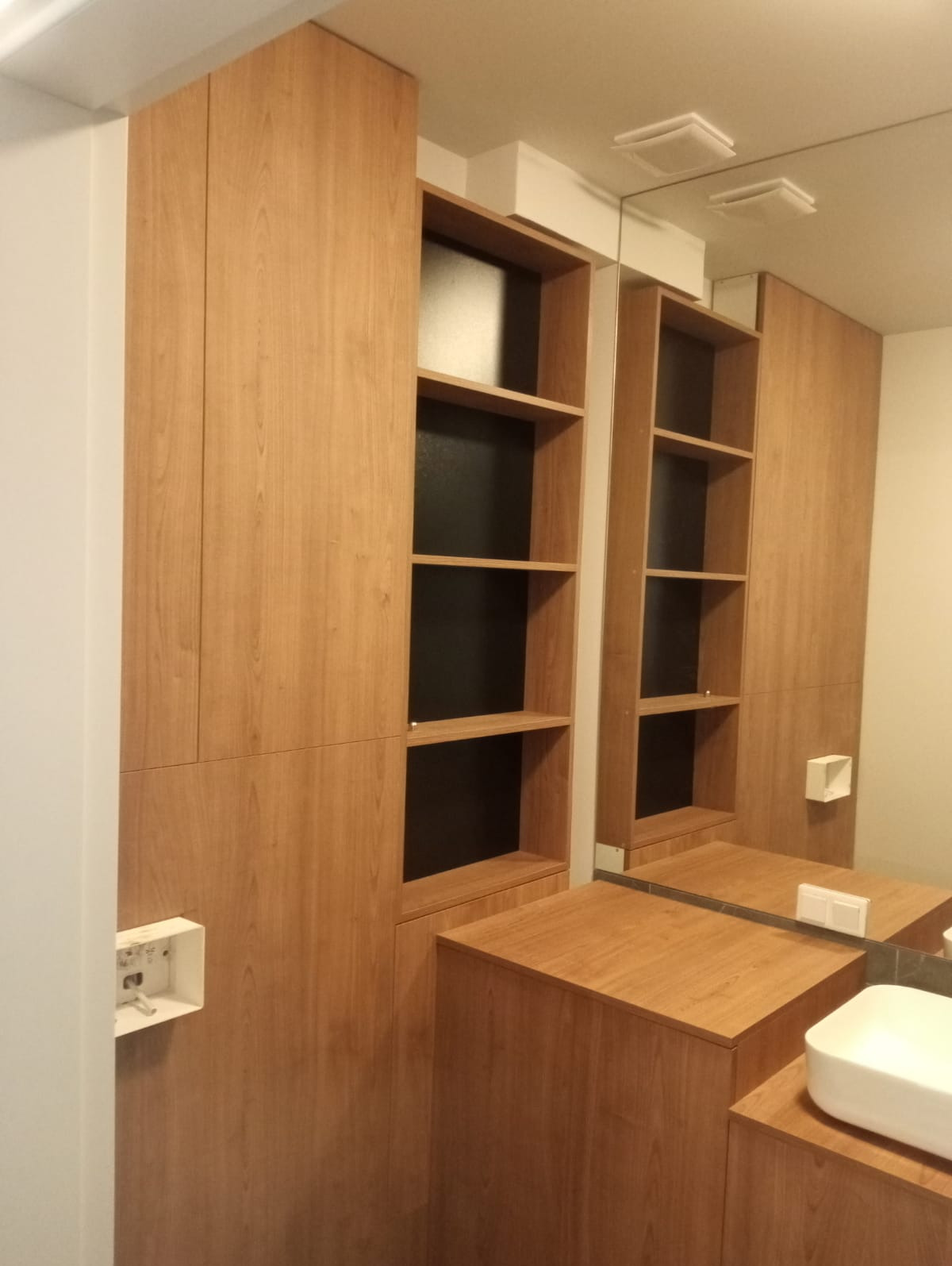 Drewniana szafa wnękowa z półkami i szafką pod umywalką, widoczne gniazdko elektryczne i odbicie w lustrze.