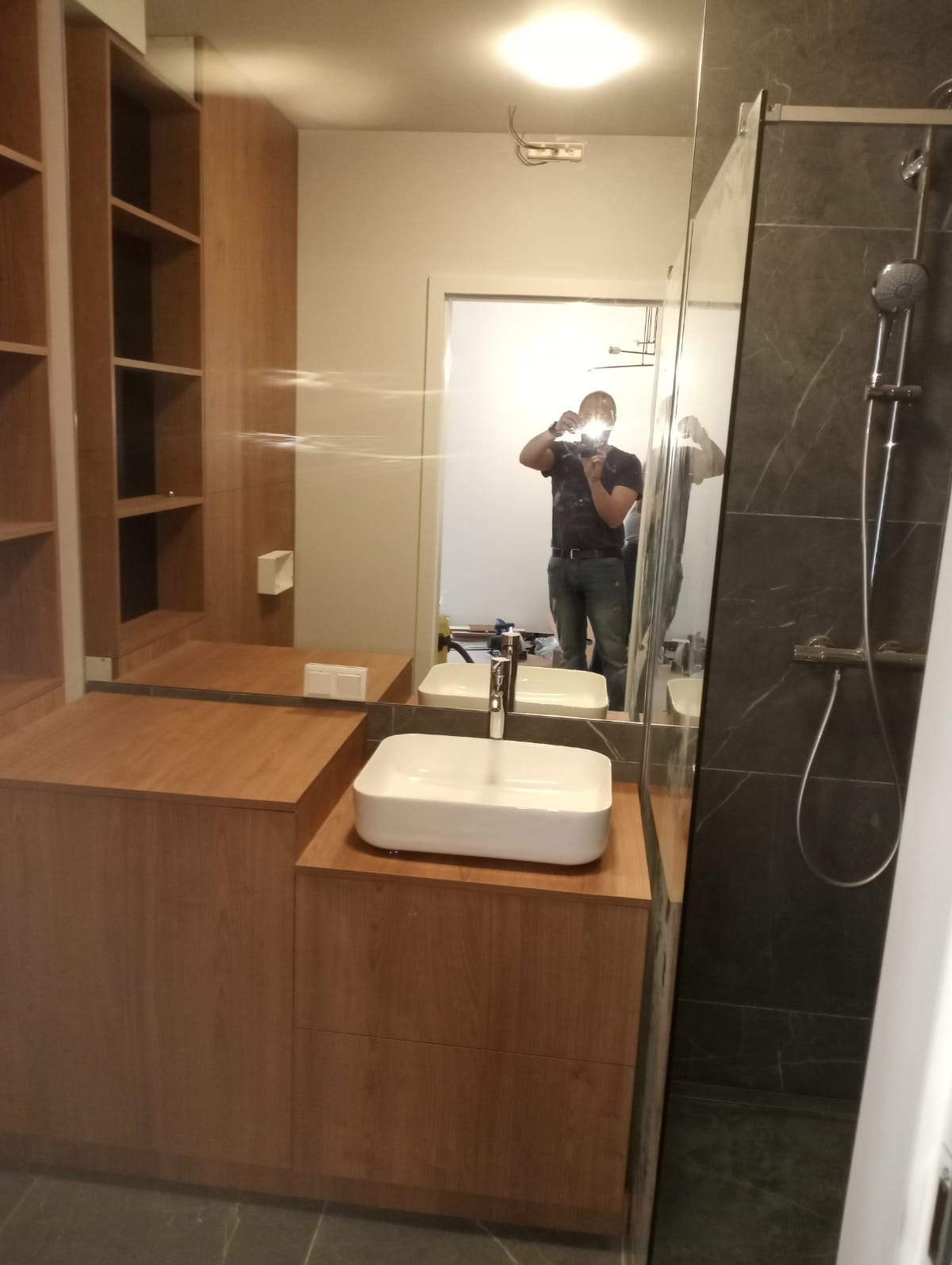 Nowoczesna łazienka z drewnianą zabudową meblową, białą umywalką nablatową i kabiną prysznicową z szarymi płytkami imitującymi marmur, widoczne odbicie osoby robiącej zdjęcie.
