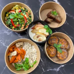 Zestaw czterech dań w ekologicznych opakowaniach: sałatka warzywna z papryką, bananowiec, jogurt z pieczonymi jabłkami i cynamonem, danie z kurczaka, ryżu i marchewki, każde udekorowane listkiem...