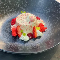 Deser panna cotta posypany czerwonym proszkiem, udekorowany truskawkami, malinami, bitą śmietaną, czerwonym biszkoptem i listkami mięty na ciemnym talerzu z efektem spękań.