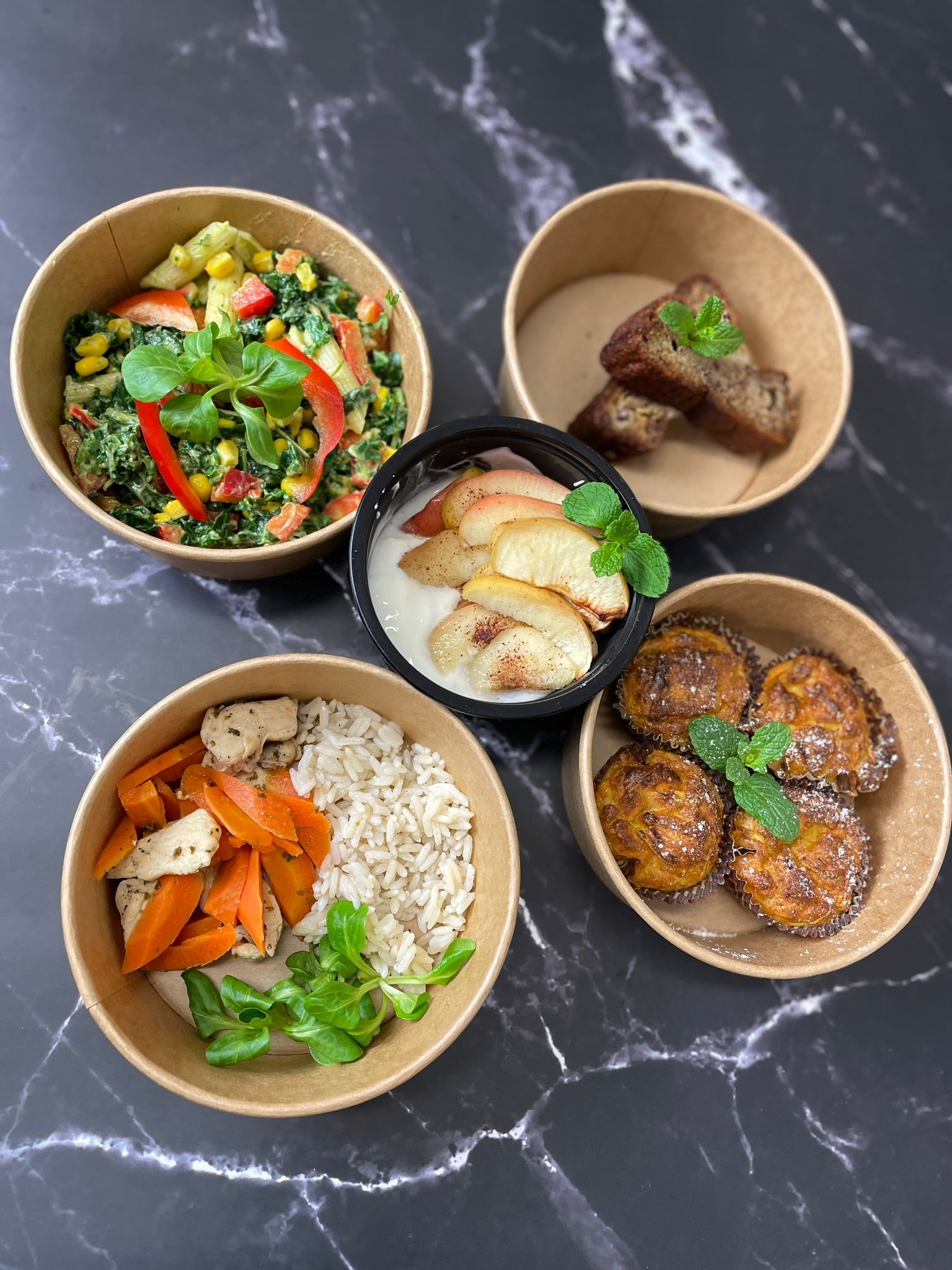 Zestaw czterech dań w ekologicznych opakowaniach: sałatka warzywna z papryką, bananowiec, jogurt z pieczonymi jabłkami i cynamonem, danie z kurczaka, ryżu i marchewki, każde udekorowane listkiem...