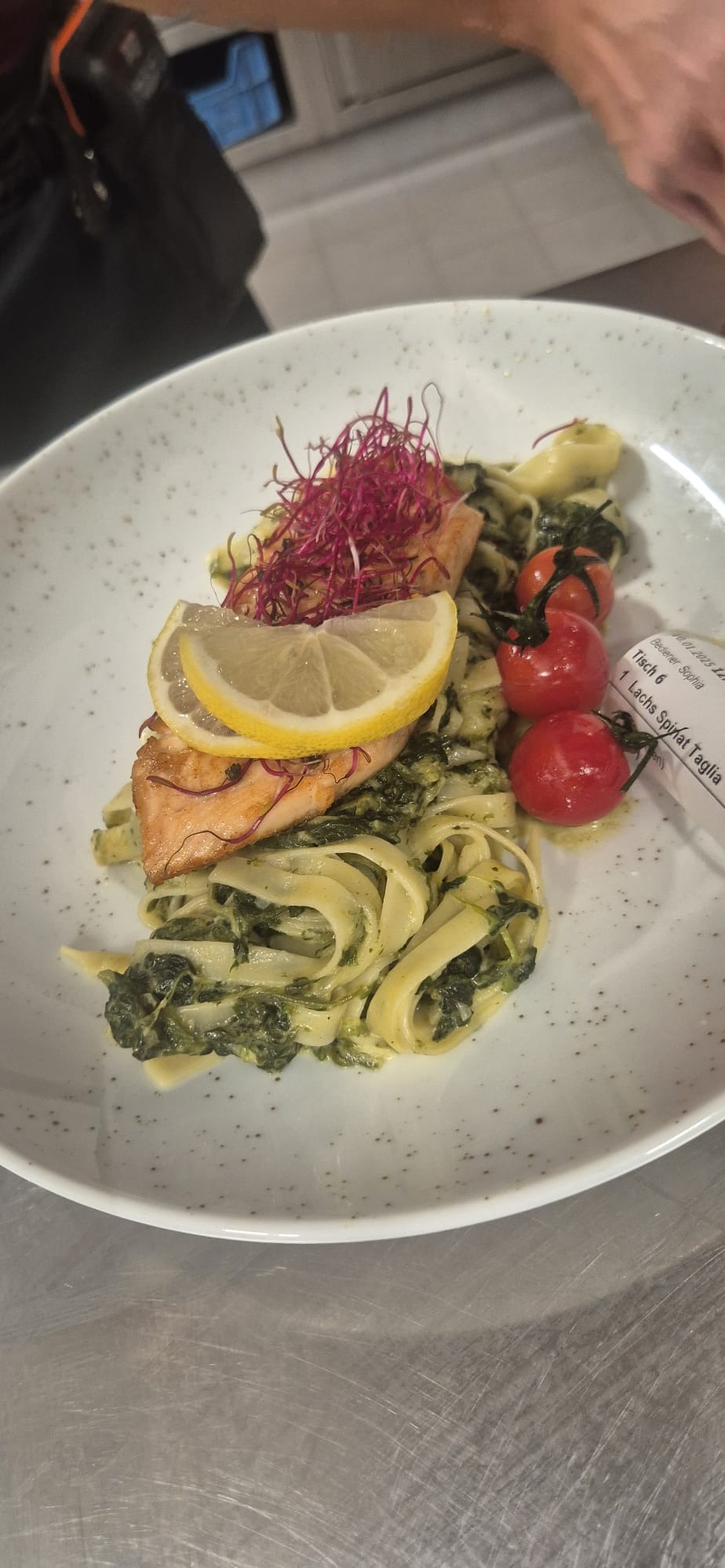 Danie z łososiem, makaronem tagliatelle ze szpinakiem, udekorowane plasterkiem cytryny, czerwoną mikro zieleniną i trzema pomidorkami cherry, podane na białym talerzu z szarymi plamkami.