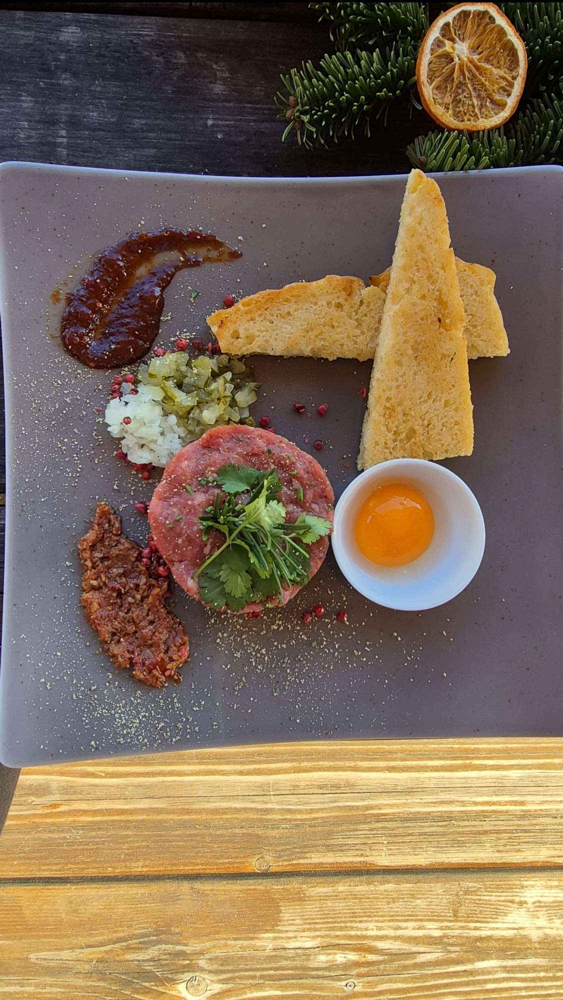 Tatar wołowy na szarym talerzu z dodatkami: sosem, ogórkiem, cebulą, grzankami i żółtkiem, ozdobiony gałązką świerku i suszoną pomarańczą.