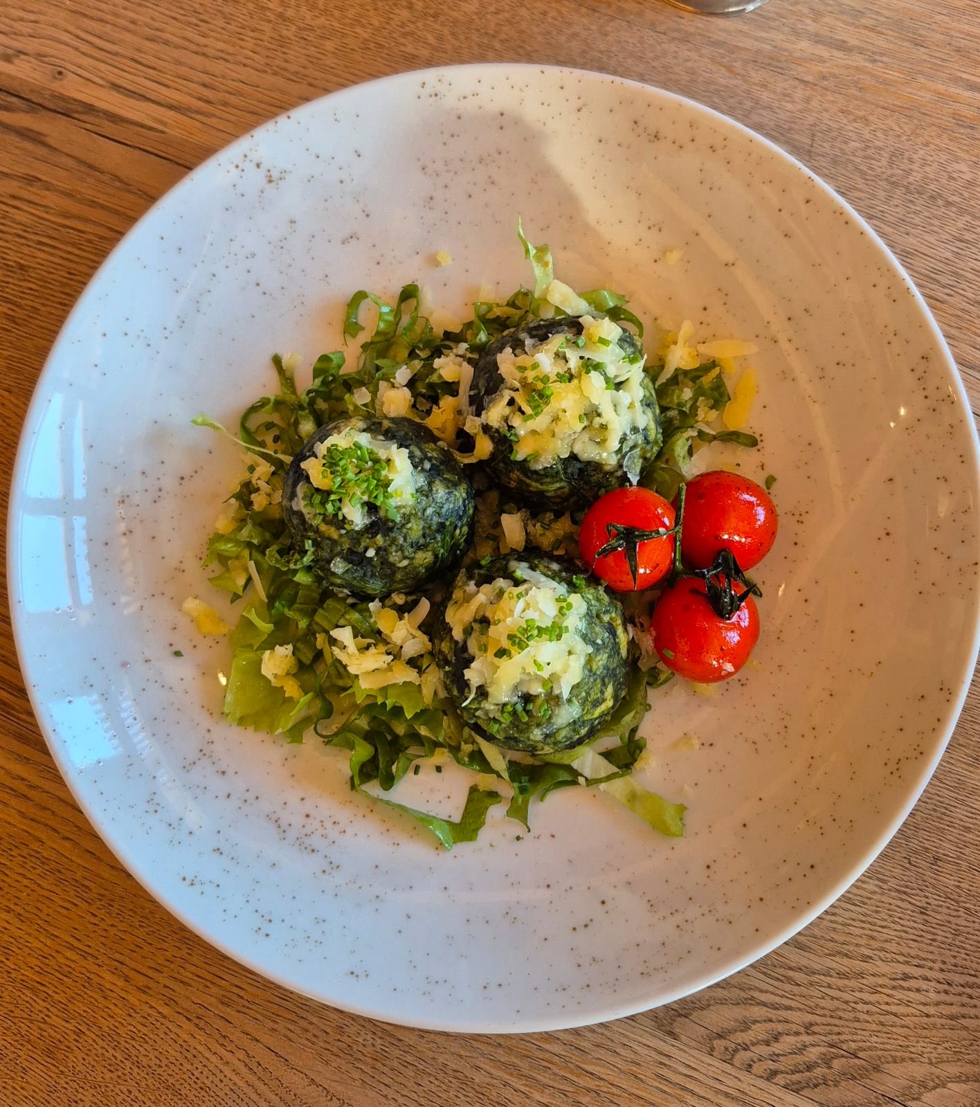 Trzy zielone kulki szpinakowe posypane serem i ziołami, ułożone na podkładzie z sałaty, obok trzy czerwone pomidorki koktajlowe, podane na białym talerzu z brązowymi plamkami, na drewnianym stole.