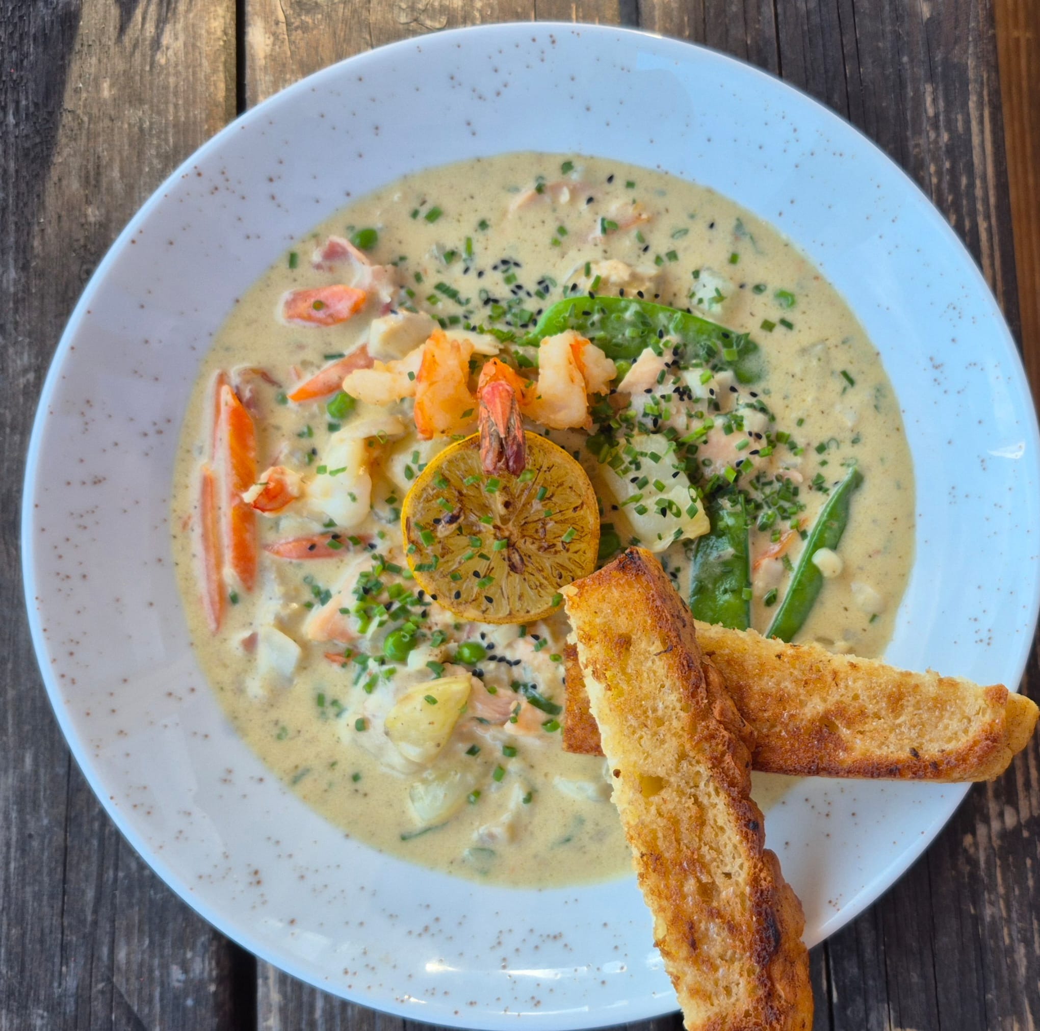 Kremowa zupa rybna z krewetkami, marchewką, groszkiem cukrowym, posypana czarnym sezamem i szczypiorkiem, podana z grillowaną cytryną i dwoma grzankami na talerzu z delikatnym wzorem.