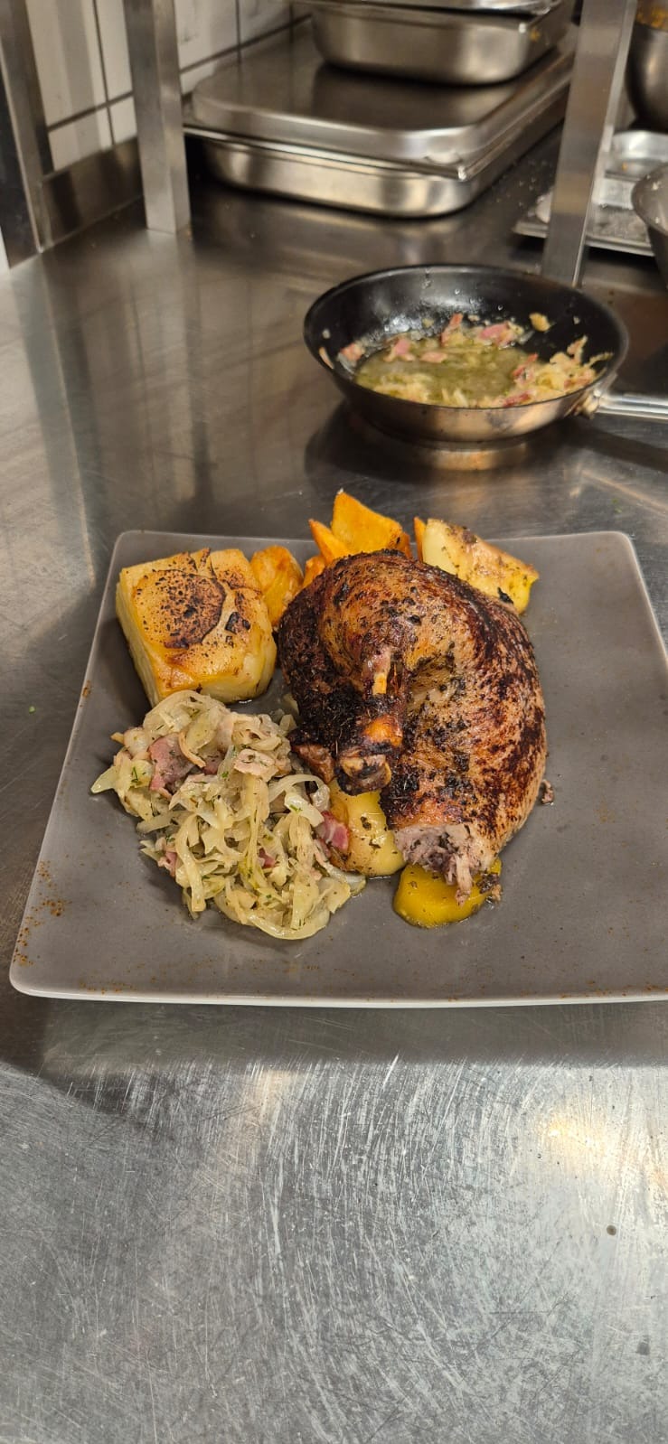 Pieczona noga z kurczaka z chrupiącą skórką, podana na szarym talerzu z pieczonymi ziemniakami, kapustą i cebulą, w tle patelnia z sosem.