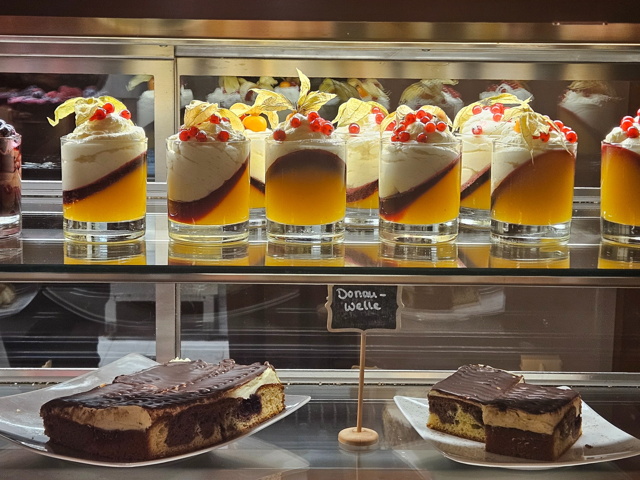 Szklana witryna chłodnicza z deserami: pucharki z kremem, galaretką i owocami oraz kawałki ciasta 'Donauwelle' na białych talerzykach.