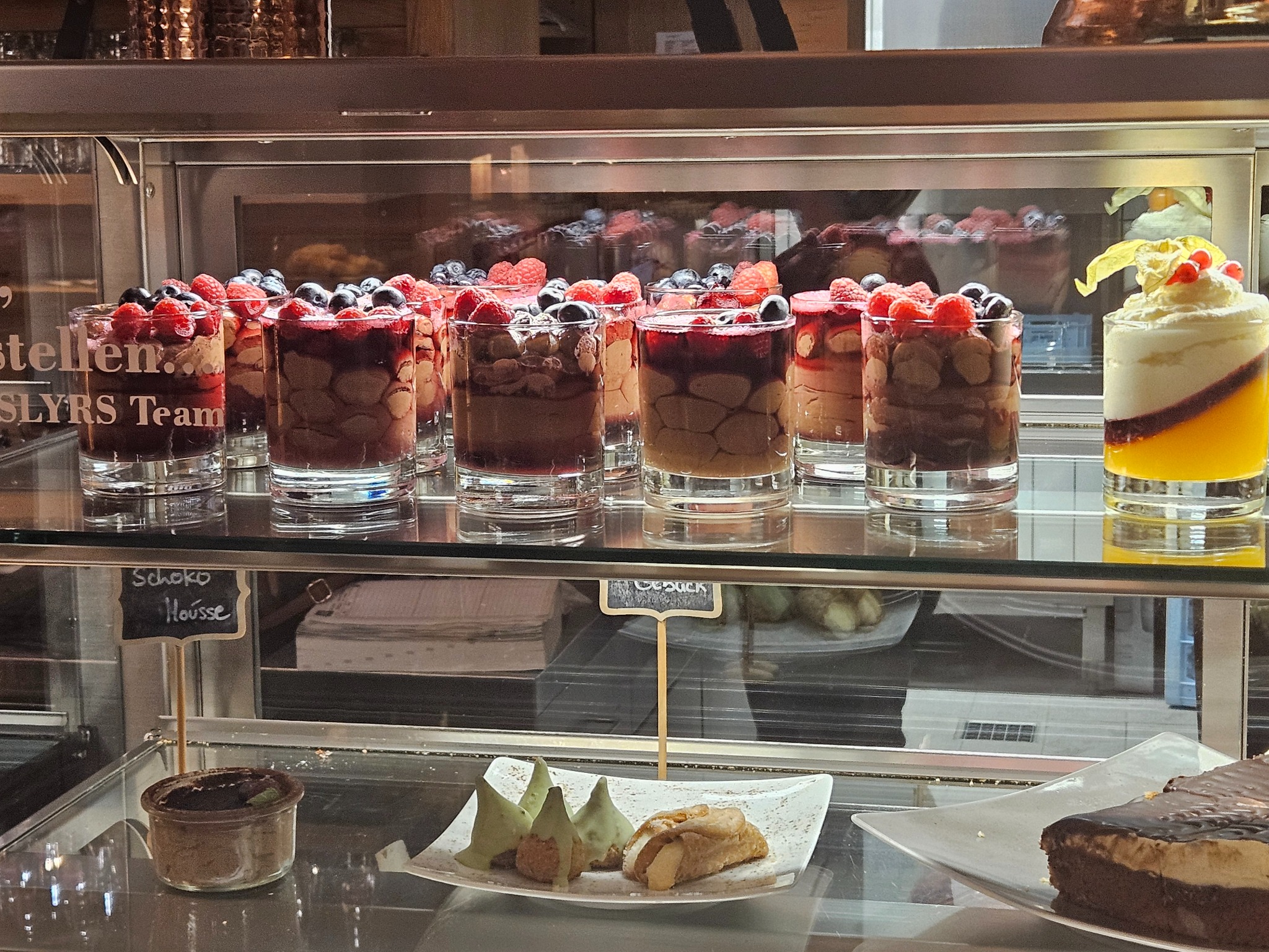 Szklana witryna chłodnicza wypełniona deserami: musy czekoladowe z owocami leśnymi w szklankach, kremowe desery z galaretką oraz ciasta na paterach.