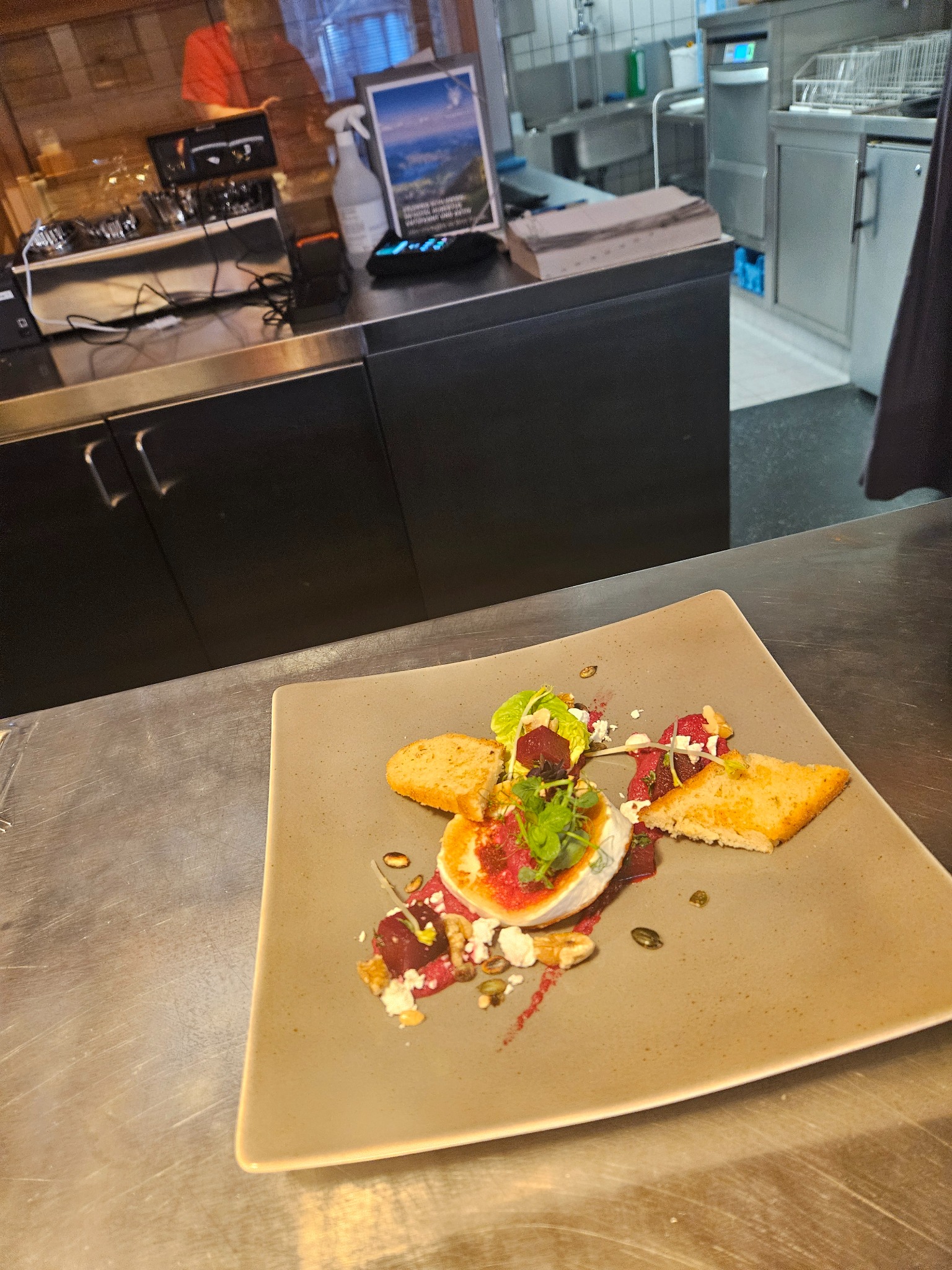 Przystawka na kwadratowym talerzu: okrągły ser kozi z burakami, sosem i grzankami, dekorowany świeżymi ziołami, na tle profesjonalnej kuchni.