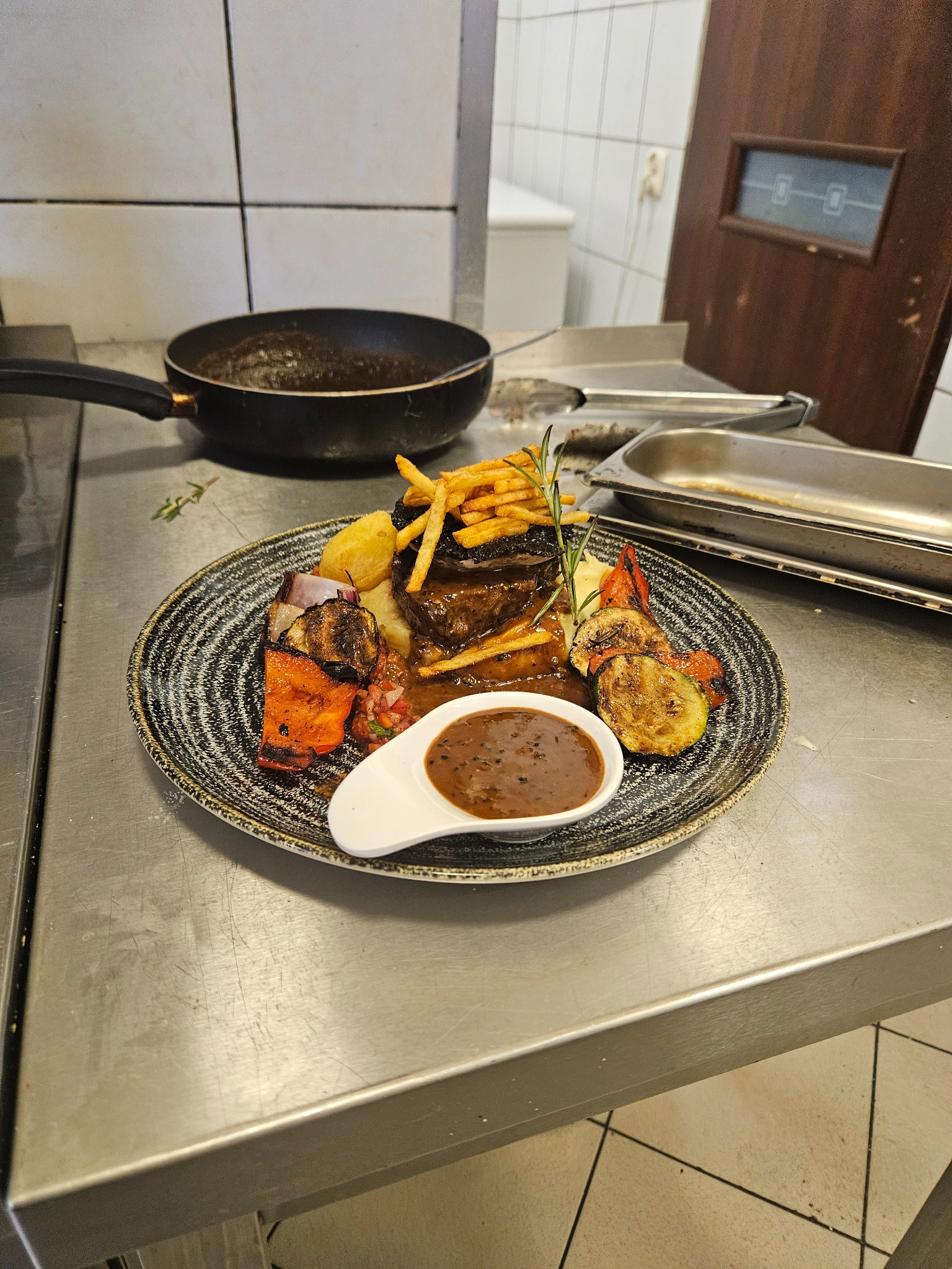 Gotowe danie: stek z frytkami, ziemniakami, grillowanymi warzywami i sosem na talerzu o wzorzystym obrzeżu, w tle patelnia, metalowy stół i drzwi.