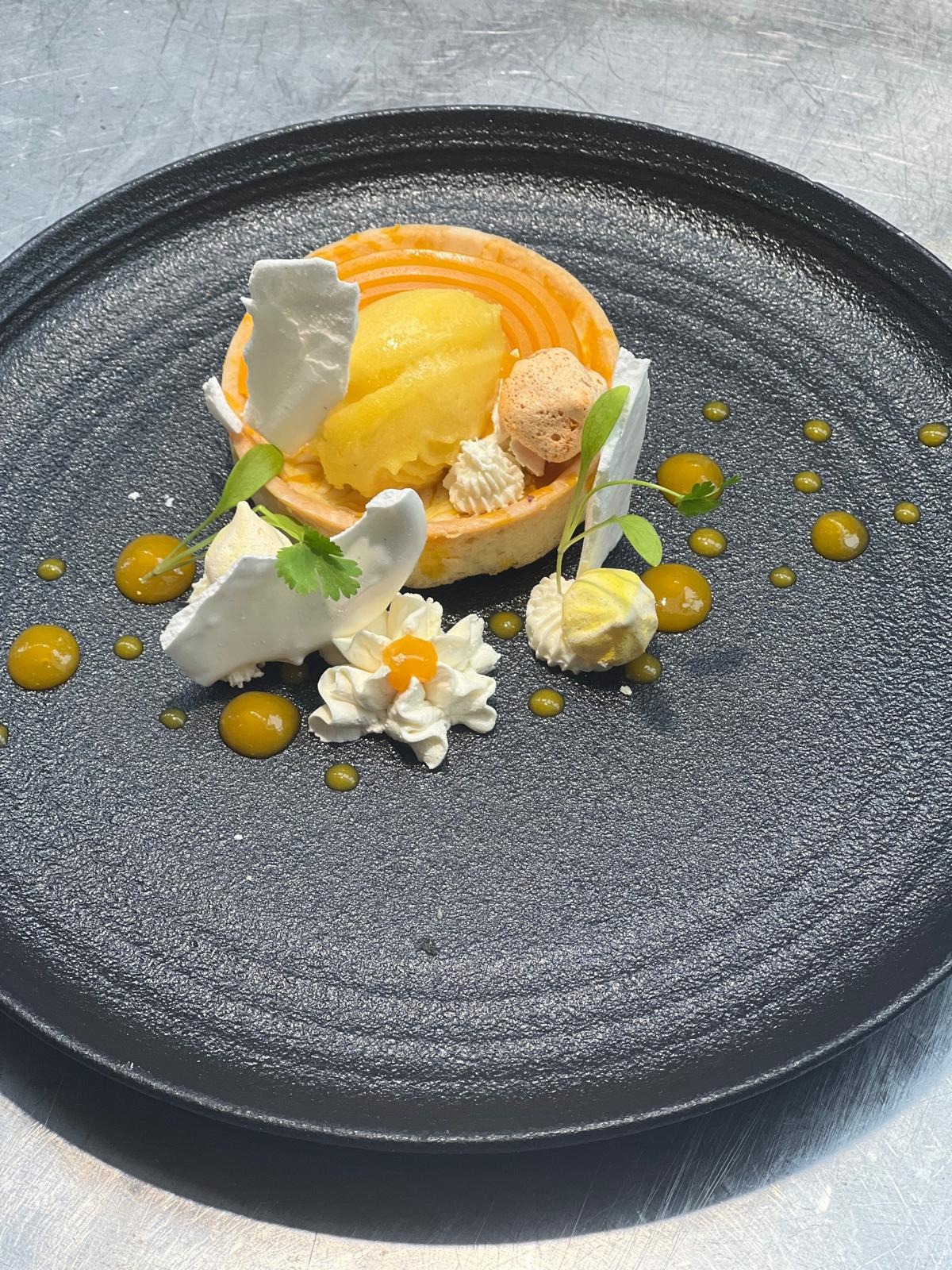 Elegancko udekorowany deser na czarnym talerzu: tarta z sorbetem mango, bezą, kremem i kroplami sosu.