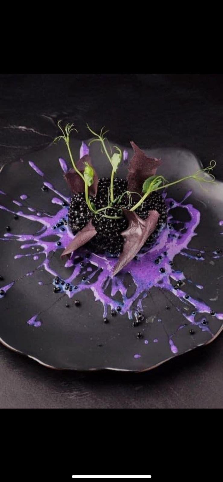 Awangardowa kompozycja deserowa na czarnym talerzu z fioletowymi akcentami, prezentowana przez cukiernię z Barczewa, z użyciem jadalnych kwiatów i czarnych kuleczek.