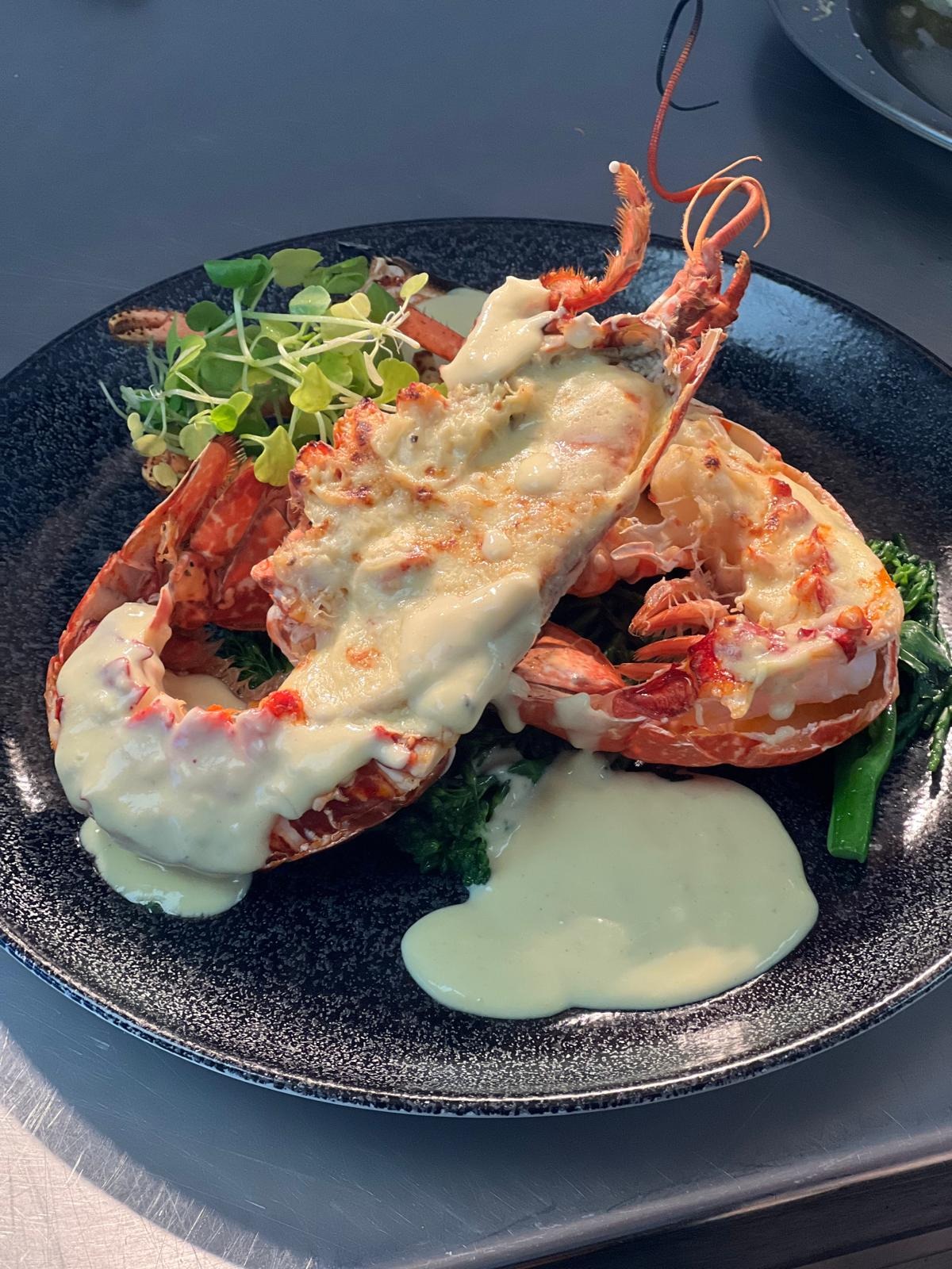 Dwa homary pod beszamelem, podane na ciemnym talerzu z brokułami i mikro-zielenią.