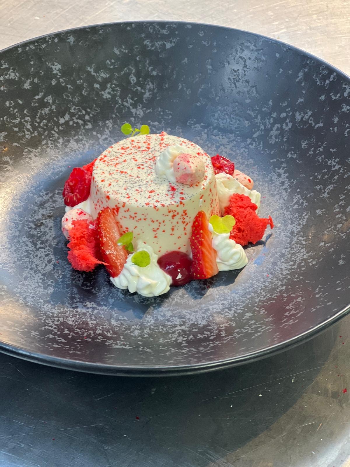 Deser panna cotta posypany czerwonym proszkiem, udekorowany truskawkami, malinami, bitą śmietaną, czerwonym biszkoptem i listkami mięty na ciemnym talerzu z efektem spękań.