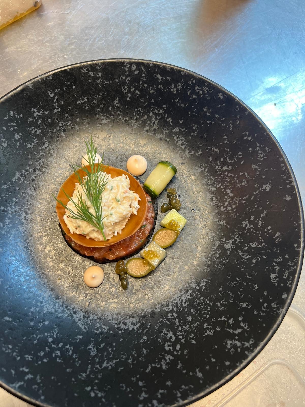 Elegancko podany tatar wołowy z kremowym sosem, dekorowany koperkiem, ogórkiem i kaparami na czarnym, nakrapianym talerzu.