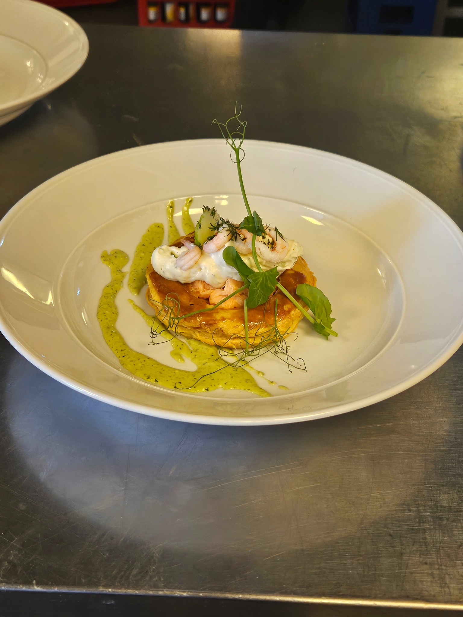 Elegancko podane danie na białym talerzu: kruche ciasto z łososiem, krewetkami, sosem śmietanowym i zielonym sosem ziołowym, udekorowane świeżymi ziołami i pędami groszku.