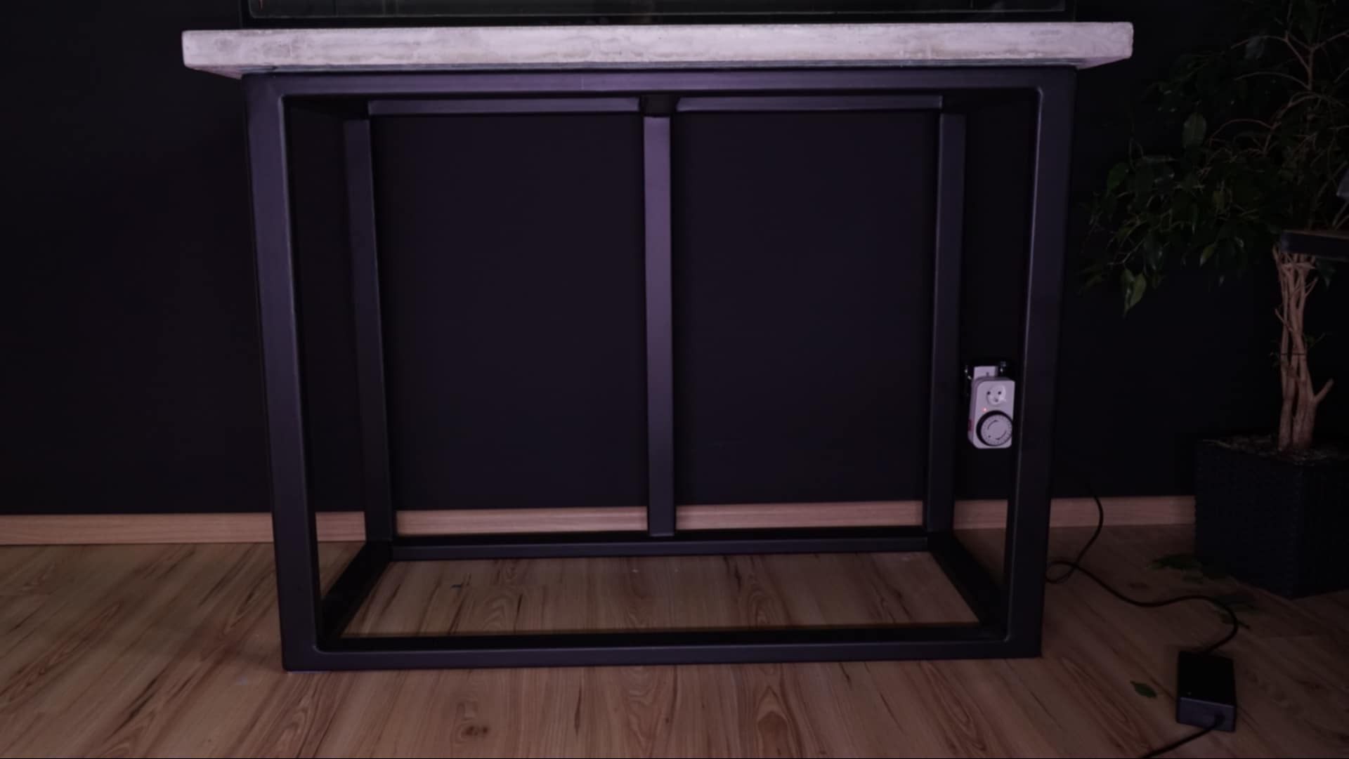 Stół z blatem imitującym beton i metalową ramą, z widocznym programatorem czasowym na nodze, stojący na drewnianej podłodze obok rośliny doniczkowej na tle ciemnej ściany.