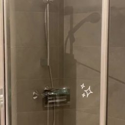 SANMIR SPÓŁKA Z OGRANICZONĄ ODPOWIEDZIALNOŚCIĄ - Szklana kabina prysznicowa z chromowaną armaturą na tle szarych płytek, widoczne ślady czyszczenia na szkle.