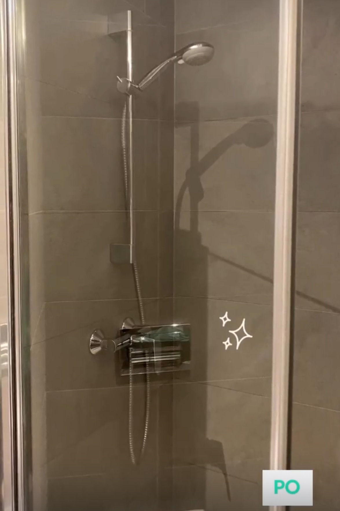 Szklana kabina prysznicowa z chromowaną armaturą na tle szarych płytek, widoczne ślady czyszczenia na szkle.