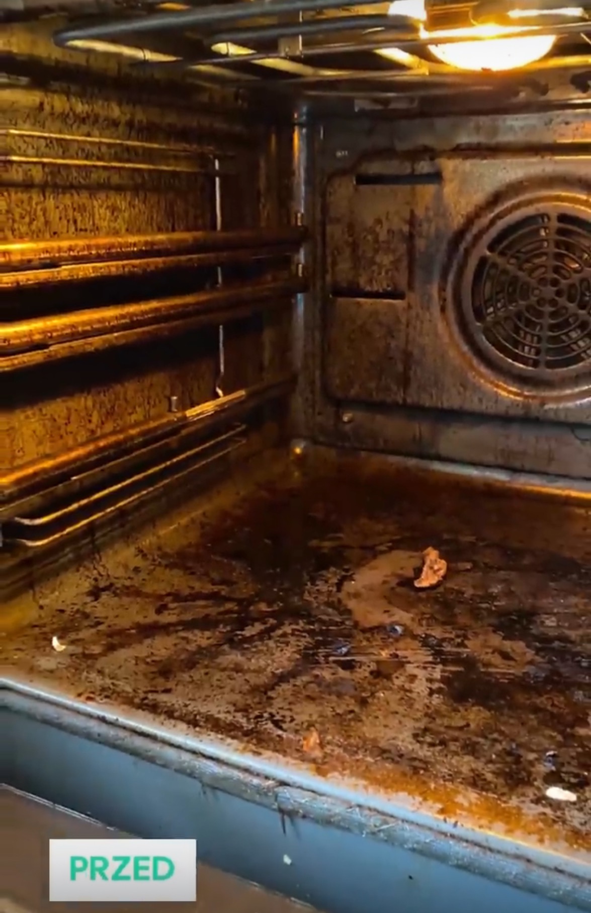 Wnętrze brudnego piekarnika przed czyszczeniem, widoczne przypalone resztki jedzenia i tłuszcz na dnie i ściankach, z włączonym oświetleniem.