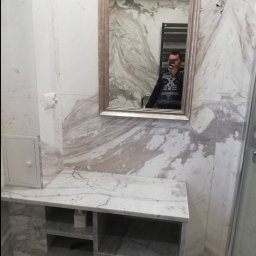 Piotr Benedyk - Wykończona łazienka z marmurowymi płytkami, widoczna szafka pod umywalkę z półkami, lustro w ramie, grzejnik drabinkowy w odbiciu lustrzanym i instalacja elektryczna do podłączenia oświetlenia.