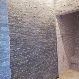 Piotr Benedyk - Ściana w łazience wyłożona szarym kamieniem dekoracyjnym, obok wejście do strefy prysznicowej z mozaiką, widoczne ślady fugowania na podłodze.