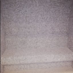 Piotr Benedyk - Wnętrze kabiny prysznicowej z siedziskiem wyłożone drobną, kwadratową mozaiką w odcieniach szarości i bieli.