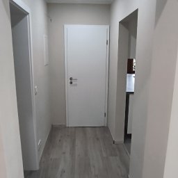 Piotr Benedyk - Korytarz po remoncie z szarą podłogą imitującą drewno, białe ściany i białe drzwi, widok w głąb mieszkania.
