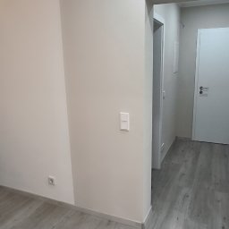 Piotr Benedyk - Wnętrze domu po remoncie: białe ściany, szare panele podłogowe, białe drzwi wewnętrzne i spiralne schody w rogu pomieszczenia.