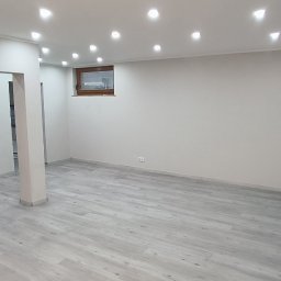 Piotr Benedyk - Wnętrze pokoju po remoncie z szarą podłogą imitującą drewno, świeżo pomalowane ściany na biało, z kominkiem z czarną obudową i dekoracyjnymi elementami, oraz oświetleniem punktowym w suficie.