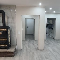 Piotr Benedyk - Wykończone wnętrze z ozdobnym piecem kaflowym na tle marmurowej ściany, szary panel podłogowy, widok na kuchnię i korytarz z białymi ścianami.