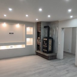 Piotr Benedyk - Jasne, przestronne wnętrze po remoncie z szarymi panelami podłogowymi, ścianą z wnękami oświetlonymi punktowo i ozdobnym piecem kaflowym na ceglanym podwyższeniu.