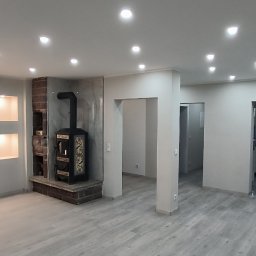 Piotr Benedyk - Przestronne, nowocześnie wykończone wnętrze z szarymi ścianami, jasną drewnopodobną podłogą, wbudowanymi półkami z oświetleniem i ozdobnym piecem kaflowym, widokiem na kuchnię w tle.