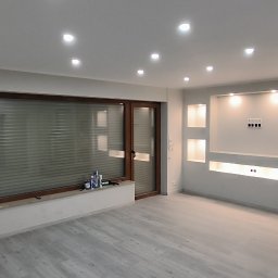 Piotr Benedyk - Wykończone wnętrze salonu z jasnymi panelami podłogowymi, ścianą z wnękami podświetlanymi LED, oknem z żaluzjami poziomymi i ozdobnym piecem kaflowym w rogu.