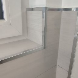 Piotr Benedyk - Wykończona narożna ściana w łazience z jasnymi płytkami imitującymi drewno i metalowymi listwami, obok okna z białą ramą.