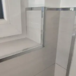 Wykończona narożna ściana w łazience z jasnymi płytkami imitującymi drewno i metalowymi listwami, obok okna z białą ramą.