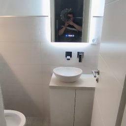 Piotr Benedyk - Nowoczesna łazienka z umywalką nablatową, podświetlanym lustrem z wyświetlaczem, minimalistyczną armaturą i płytkami imitującymi drewno na podłodze. W odbiciu lustra widoczna osoba robiąca zdjęcie.