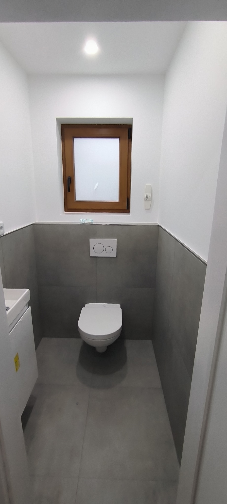 Nowoczesna, minimalistyczna łazienka z szarymi płytkami, białym sedesem i drewnianym oknem, oświetlona punktowym światłem.