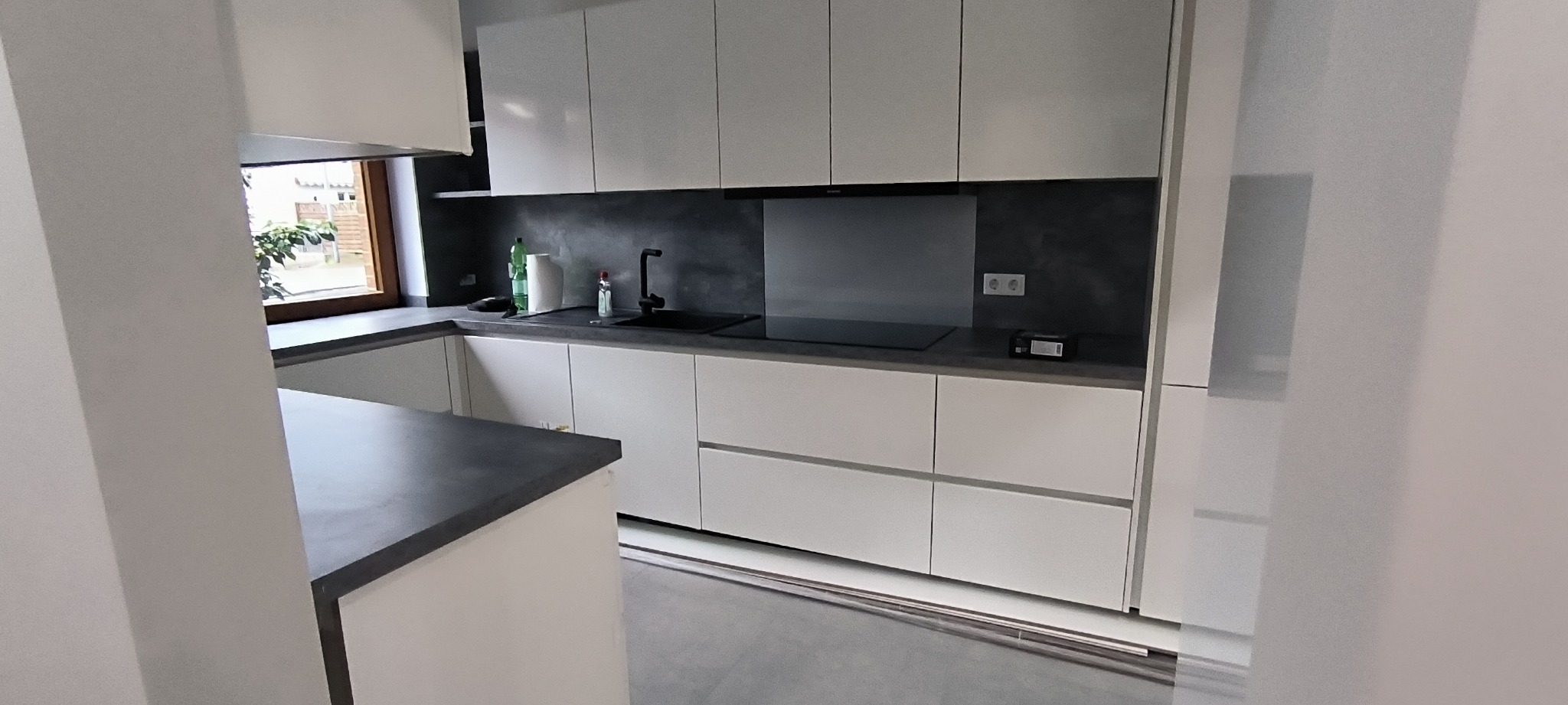 Nowoczesna, biała kuchnia z szarym blatem, czarną armaturą i szklanym panelem nad płytą grzewczą, widok z boku na wyspę kuchenną, okno z widokiem na zieleń za nim.