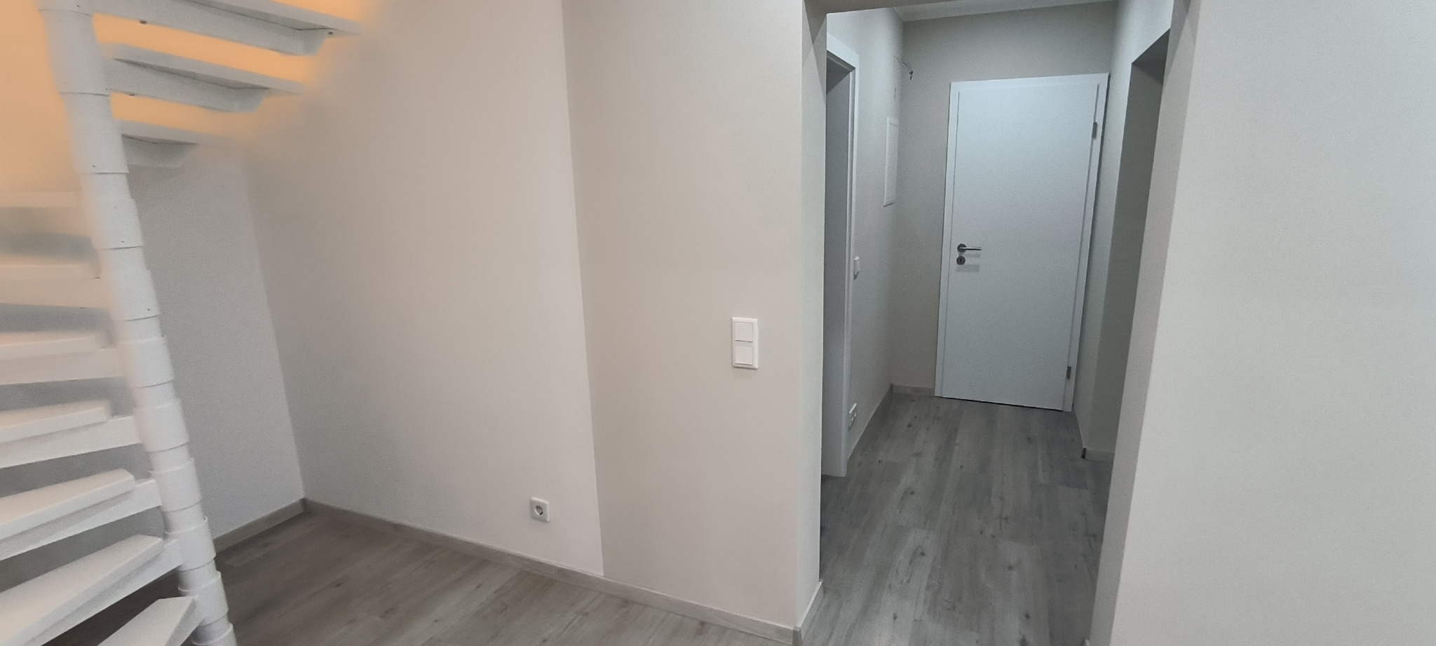 Wnętrze domu po remoncie: białe ściany, szare panele podłogowe, białe drzwi wewnętrzne i spiralne schody w rogu pomieszczenia.