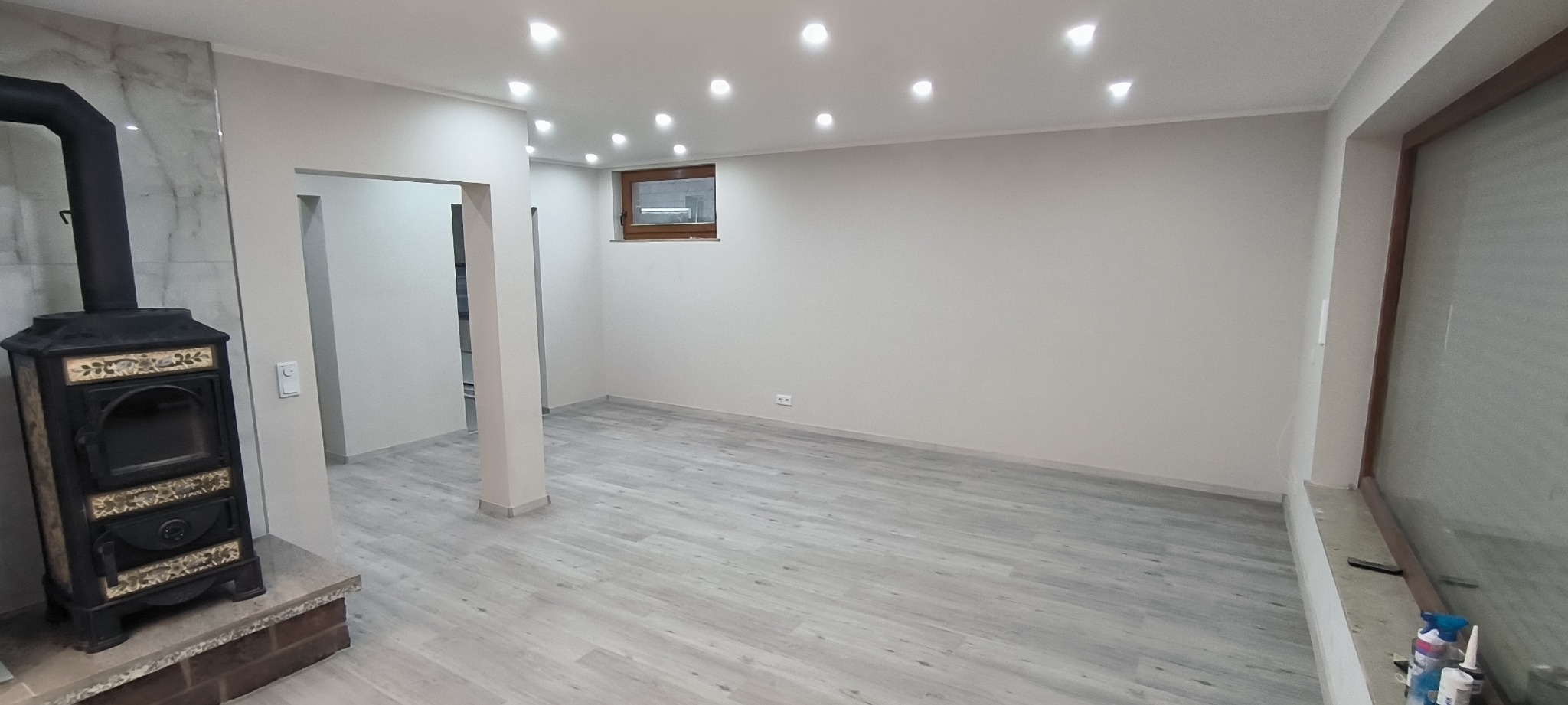 Wnętrze pokoju po remoncie z szarą podłogą imitującą drewno, świeżo pomalowane ściany na biało, z kominkiem z czarną obudową i dekoracyjnymi elementami, oraz oświetleniem punktowym w suficie.