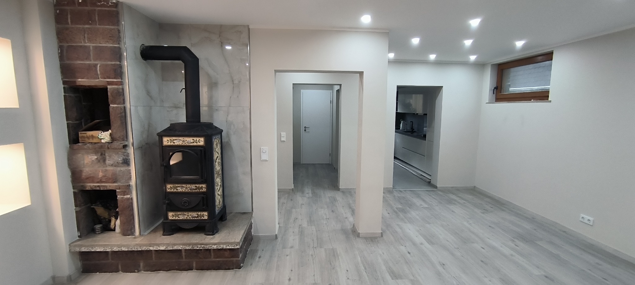Wykończone wnętrze z ozdobnym piecem kaflowym na tle marmurowej ściany, szary panel podłogowy, widok na kuchnię i korytarz z białymi ścianami.