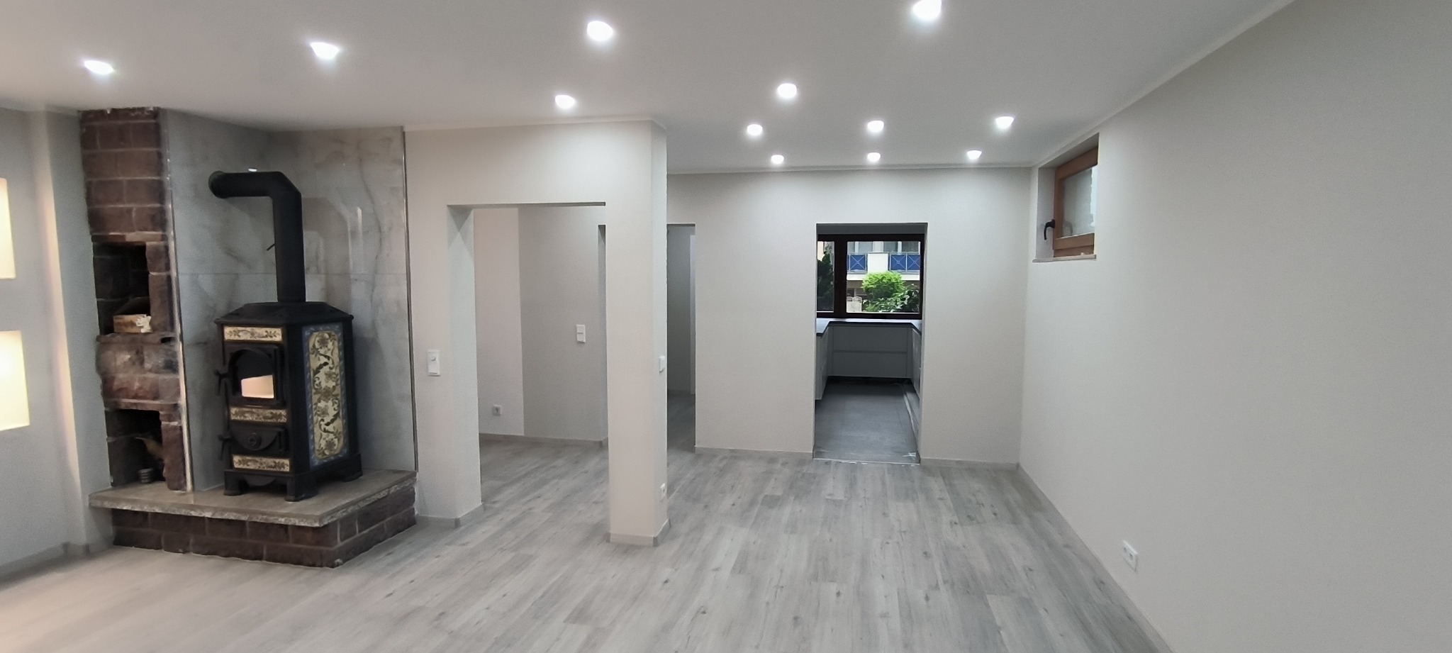 Jasne, przestronne wnętrze po remoncie z drewnopodobną podłogą, kominkiem kaflowym z ceglaną obudową i widokiem na kuchnię przez otwartą przestrzeń, oświetlone punktowymi lampami sufitowymi.
