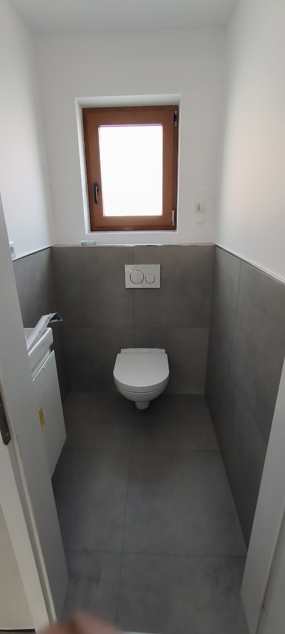 Nowoczesna toaleta w szaro-białej kolorystyce z drewnianym oknem, widoczna podwieszana miska WC z przyciskiem spłukującym na ścianie, szare płytki na ścianach i podłodze, częściowo widoczna...