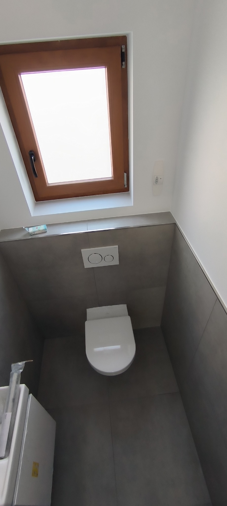 Nowoczesna łazienka z szarą glazurą, podwieszana toaleta z przyciskiem spłukującym i drewniane okno dachowe w minimalistycznym stylu.