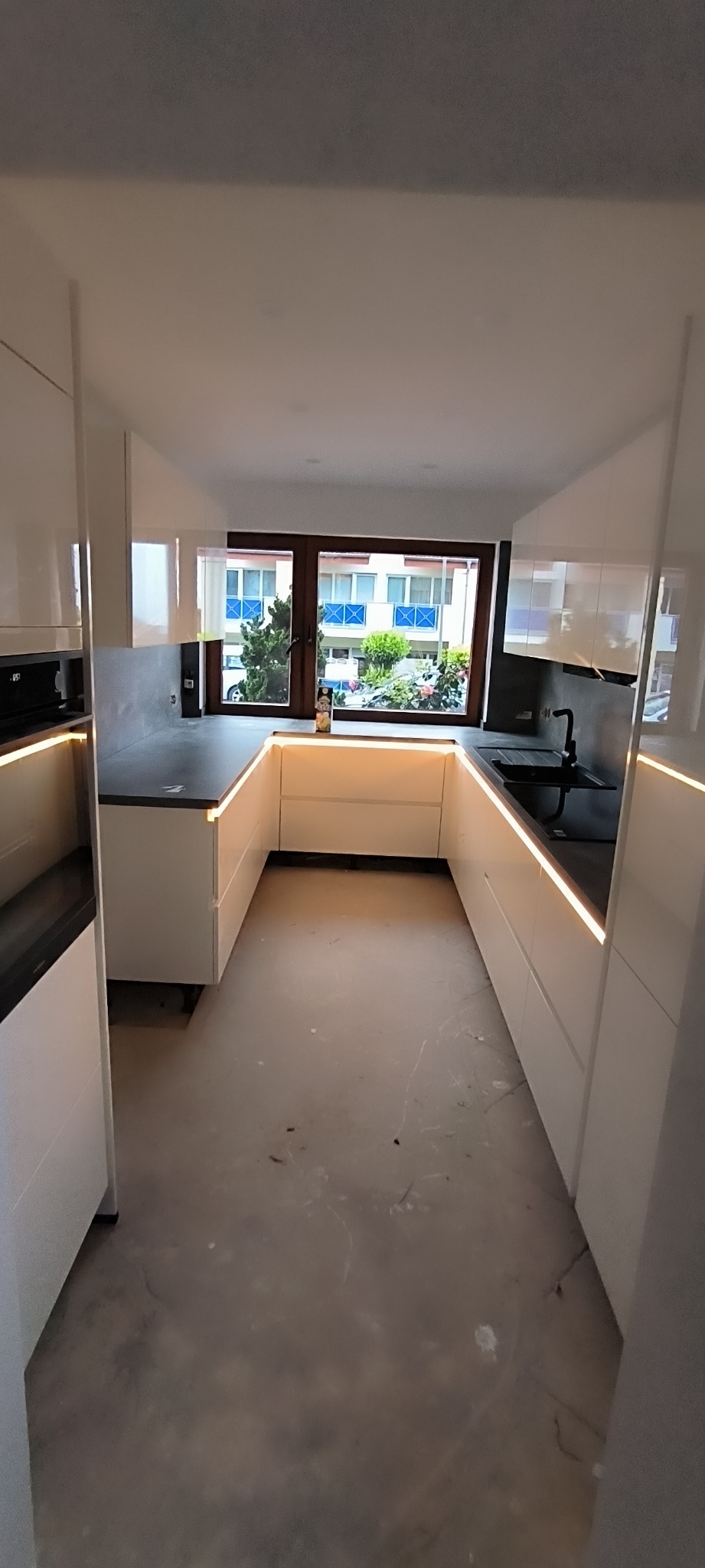 Nowoczesna, biała kuchnia w kształcie litery U z podświetleniem LED pod szafkami wiszącymi i blatem, widok na okno z zielenią na zewnątrz, surowa podłoga w trakcie remontu.