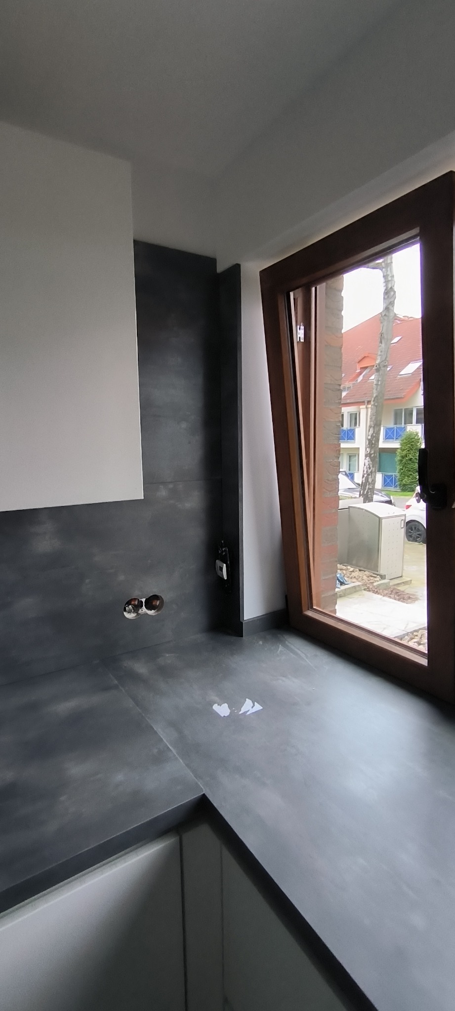 Fragment nowej, surowej łazienki z szarą, betonową okładziną ścian i podłogi, widoczne wystające kable elektryczne i fragment okna z drewnianą ramą.